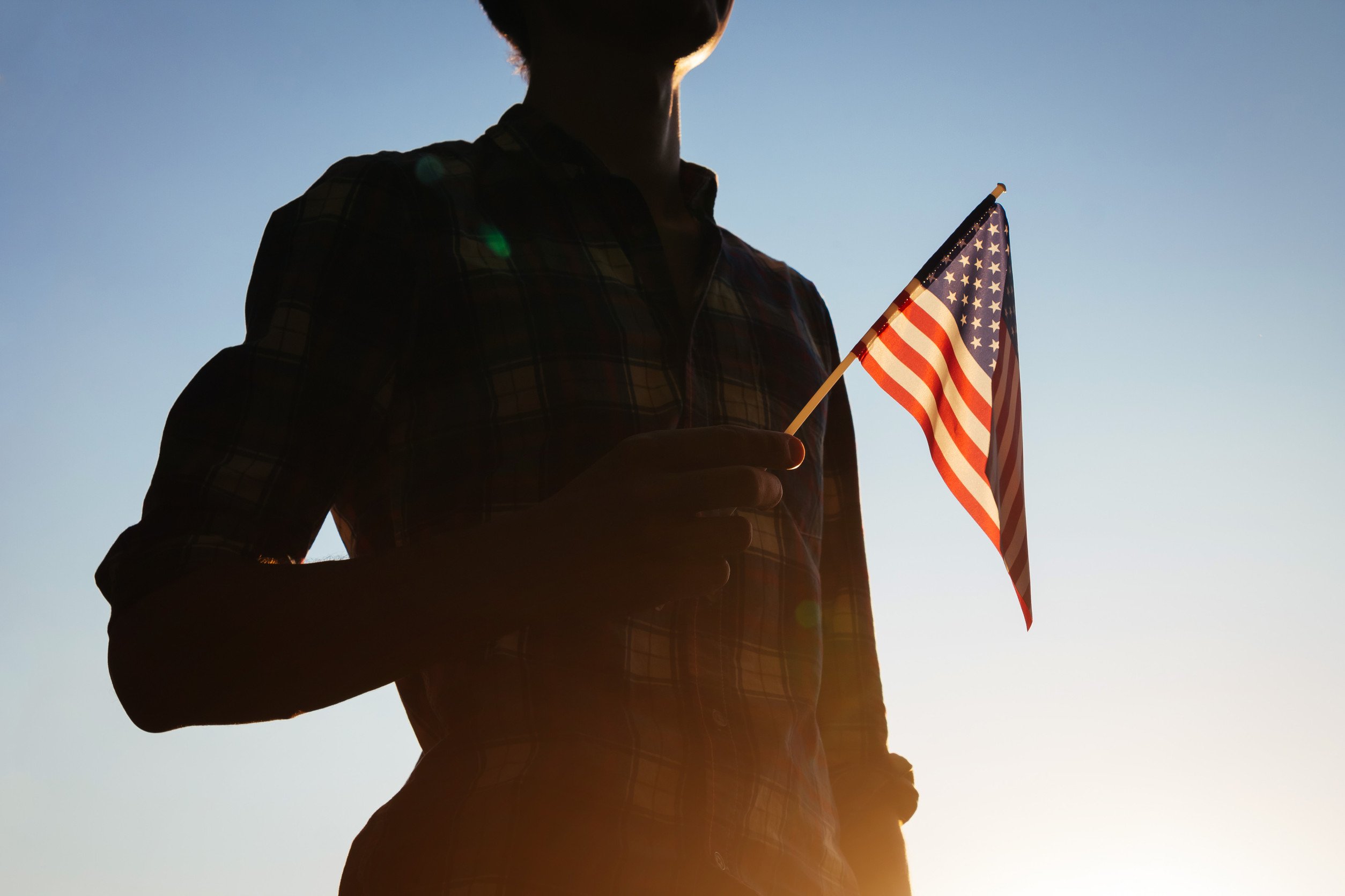 a military veteran stands holding a small american flag in silhouette against a pale blue sky