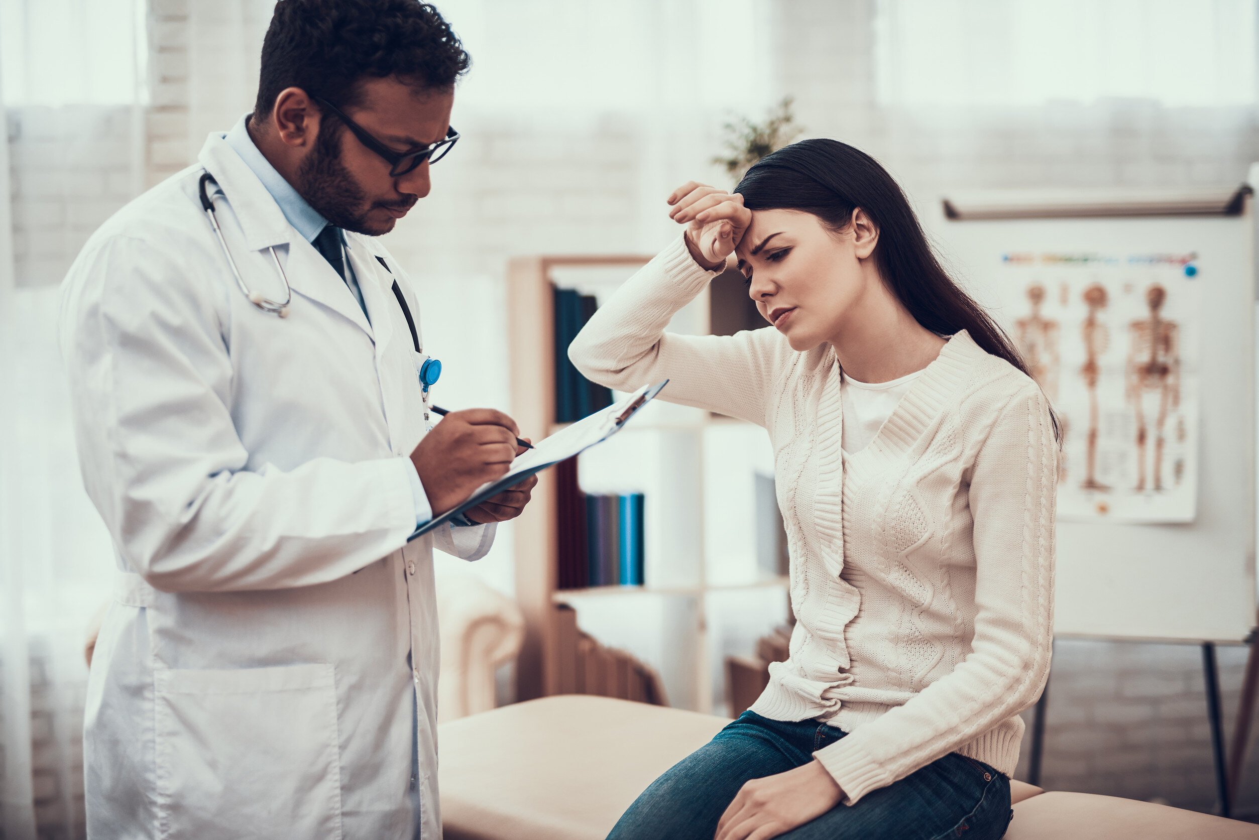 a doctor holding a clipboard takes notes on a patient holding the back of her hand against her forehead