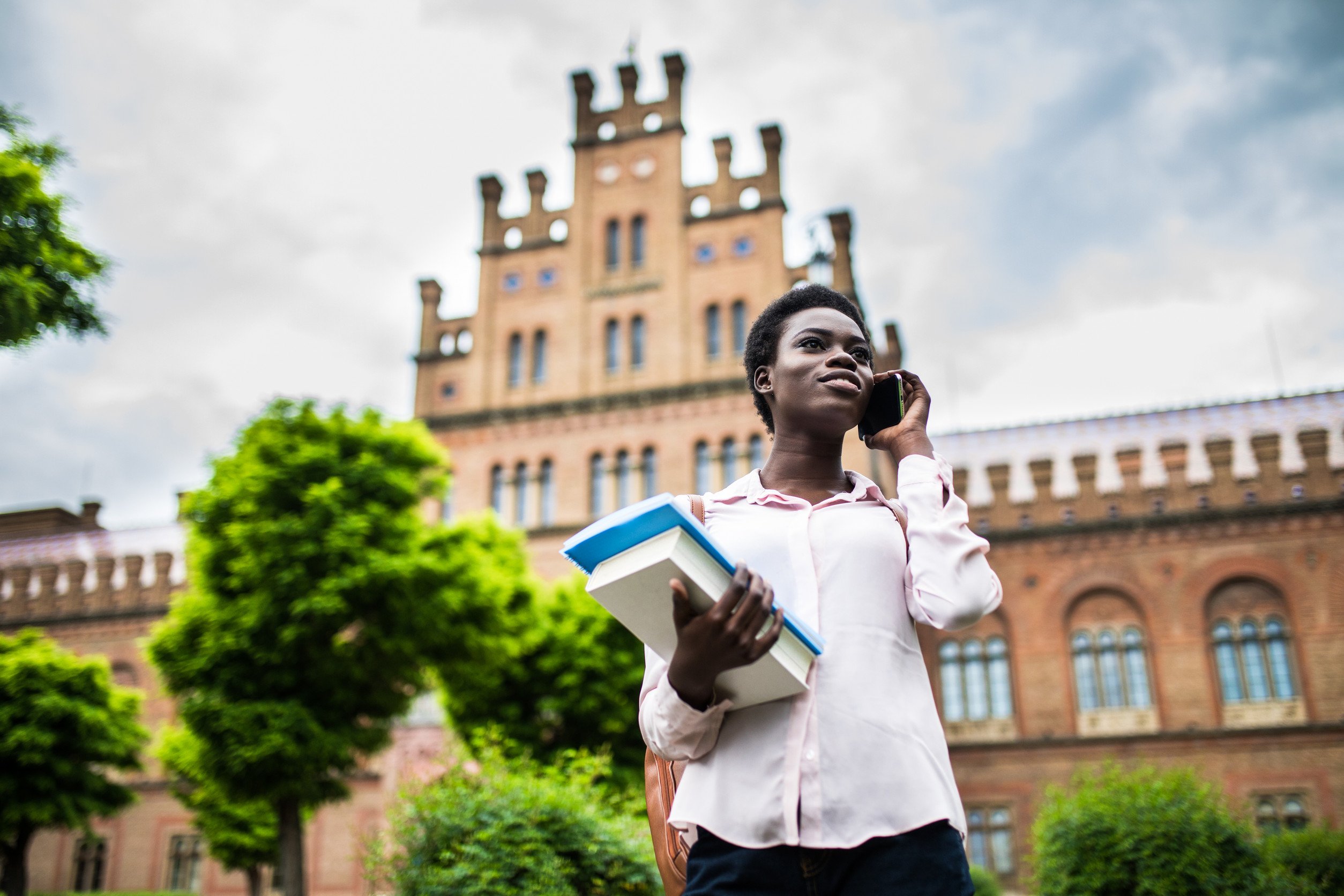 a woman holding a cell phone in one hand and textbooks in the other stands before a university building