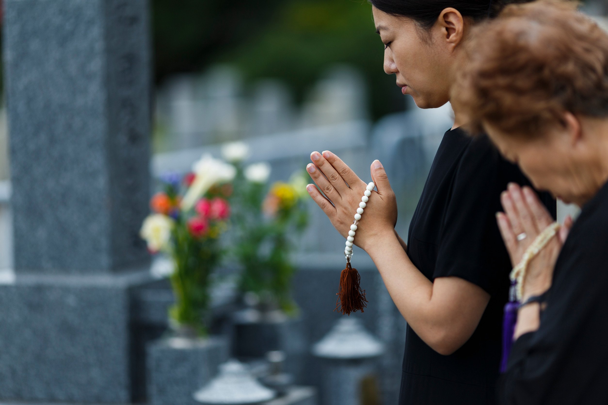 two women with their hands closed in prayer pay their respects before a gravestone