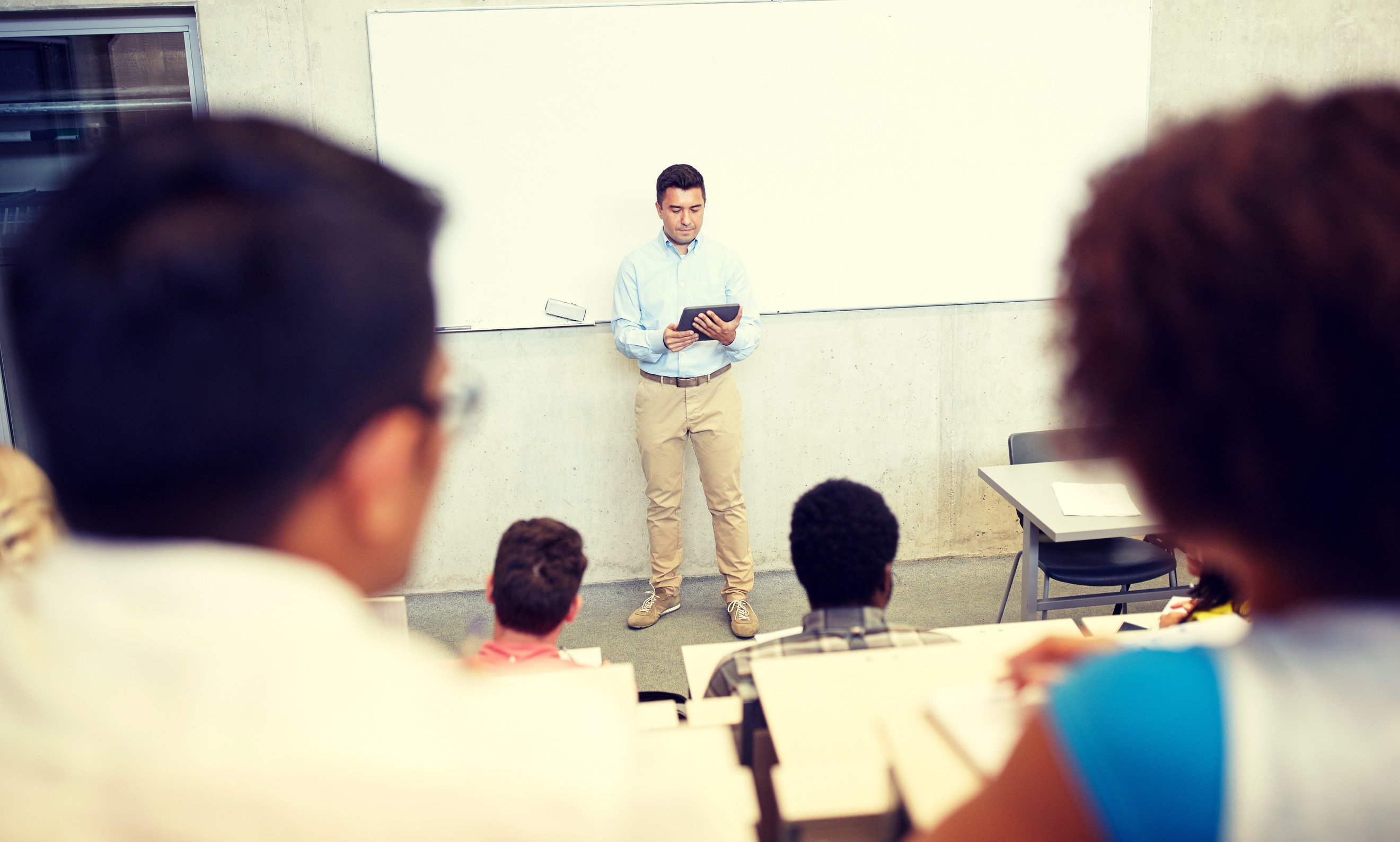 a college professor standing at the front of a class of students with a computer tablet in hand