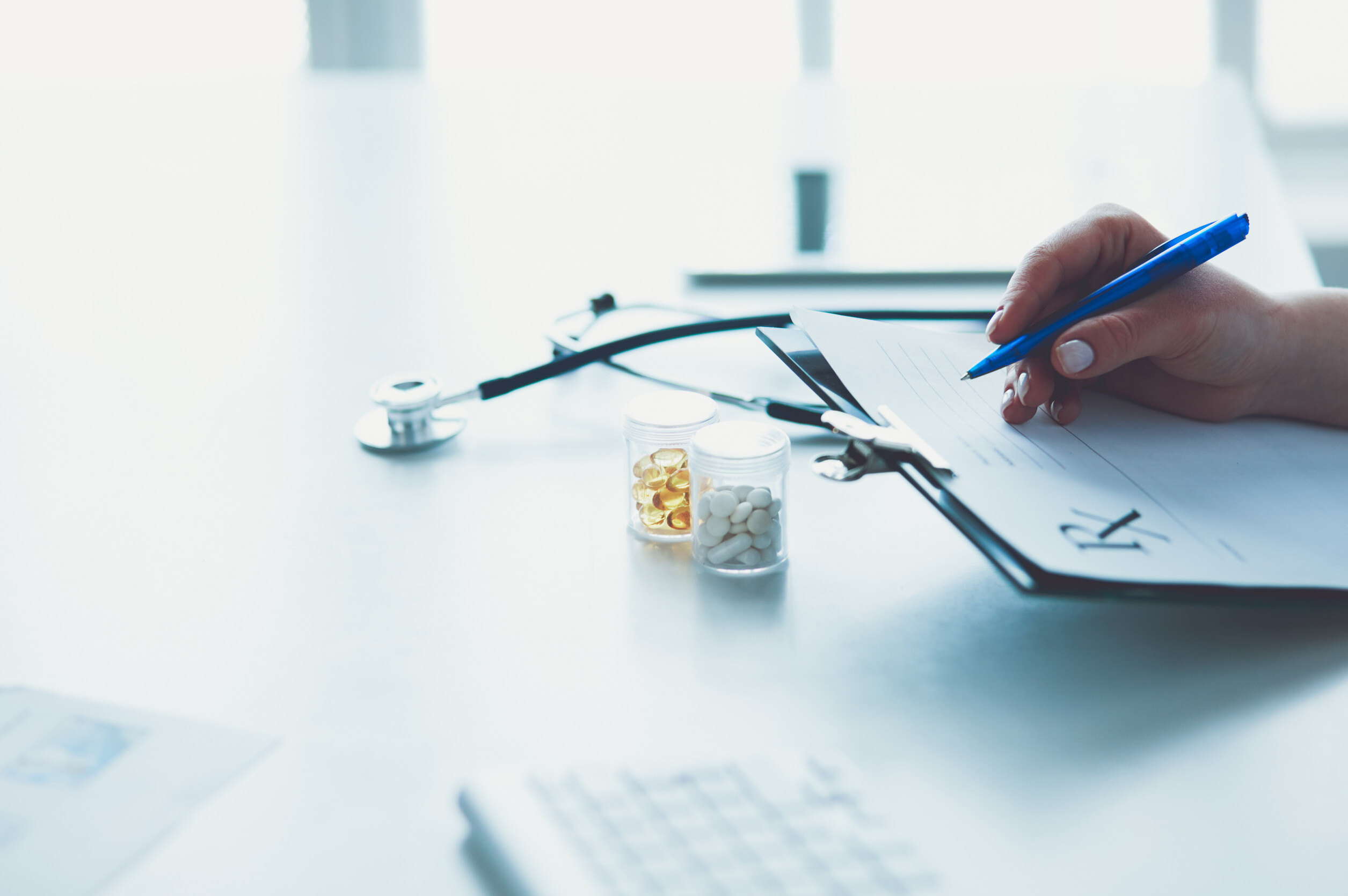 a doctor writes on a clipboard over a desk with a stephascope and pill bottles on it