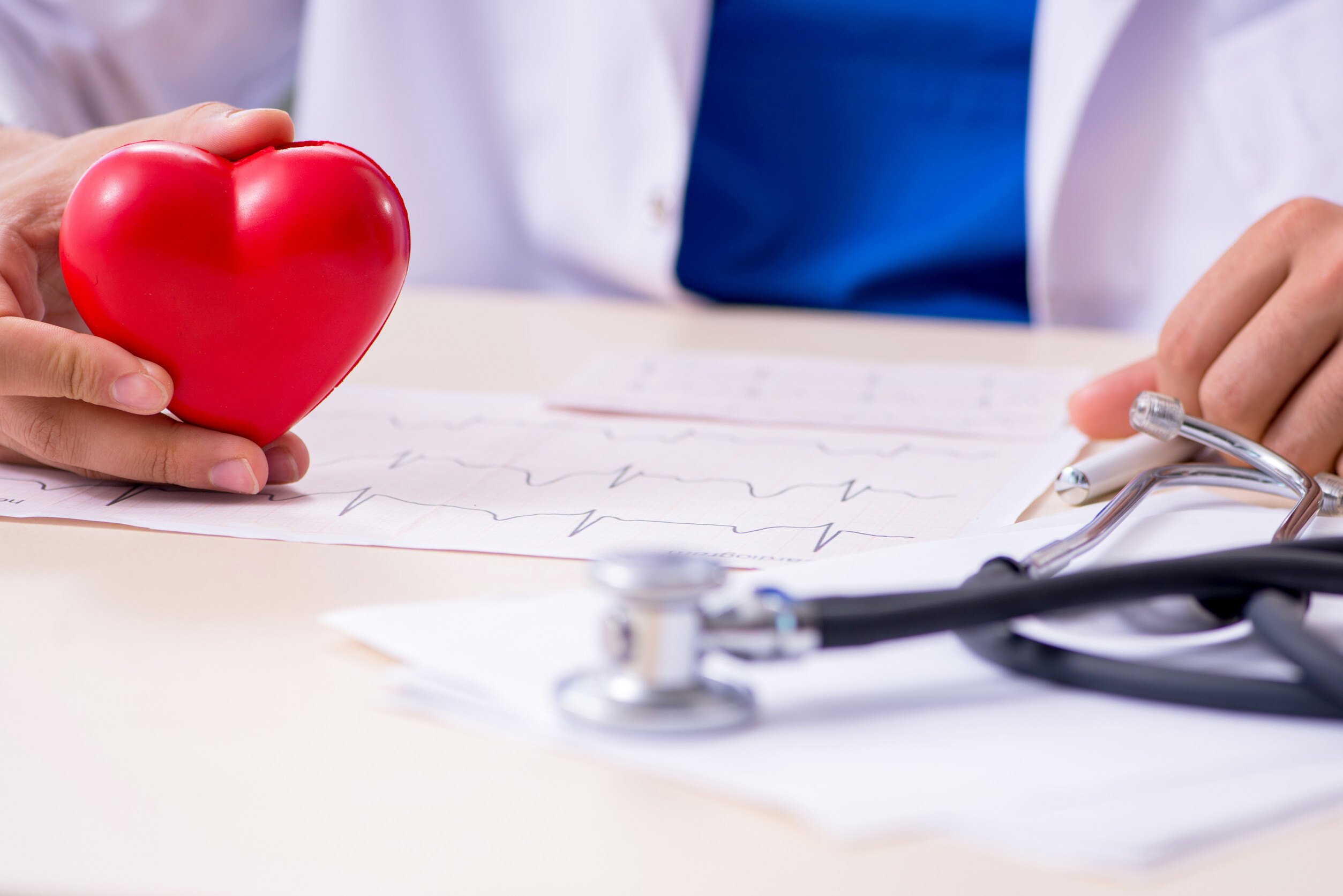 a doctor sits at their desk holding a figure of a heart while looking at ekg charts