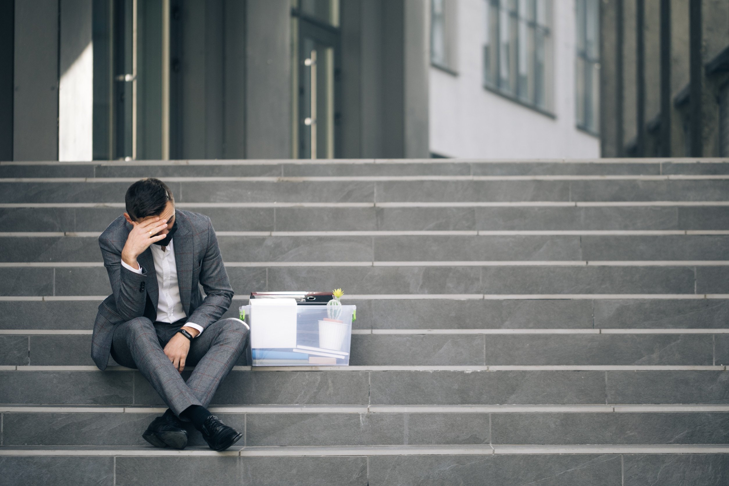 laid off employee sitting on the steps outside of his workplace with a box of personal belongings beside him