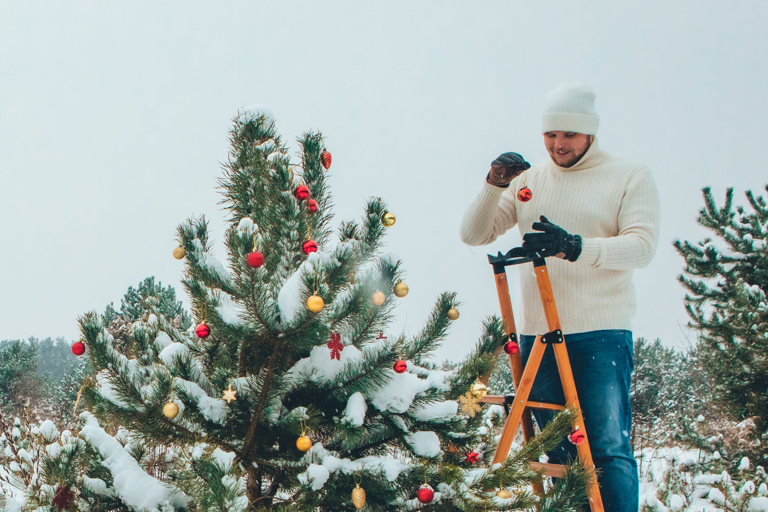 a man stands on a ladder to decorate the top of a christmas tree outside in the snow
