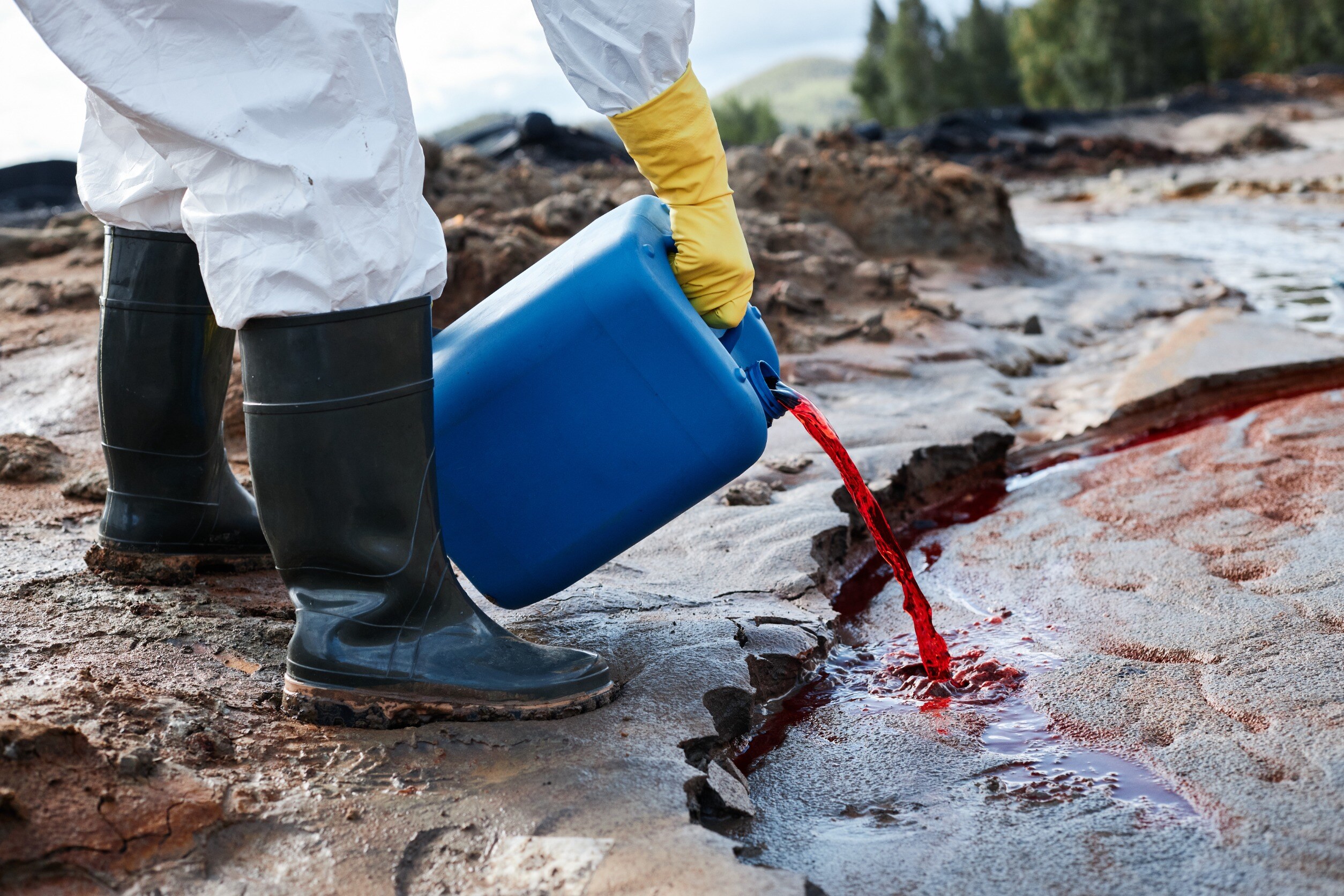 A person in a white protective suit and rubber boots pours liquid substance from a blue can into natural environment