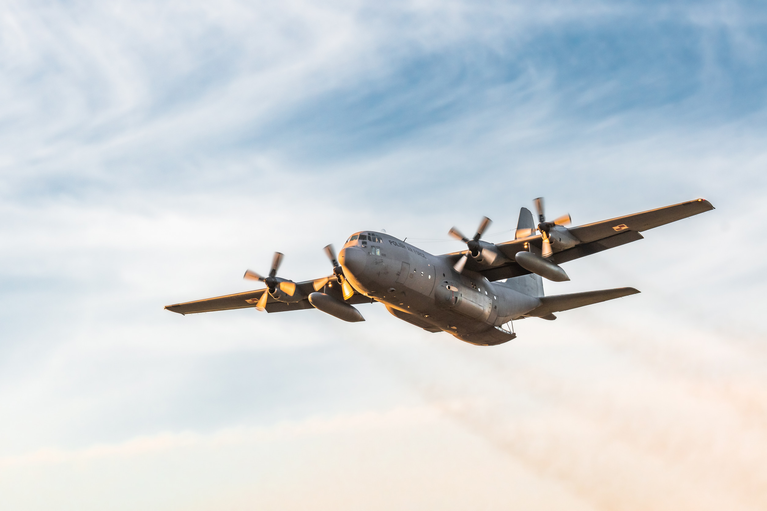 a military cargo plane flies through a blue sky and wispy clouds