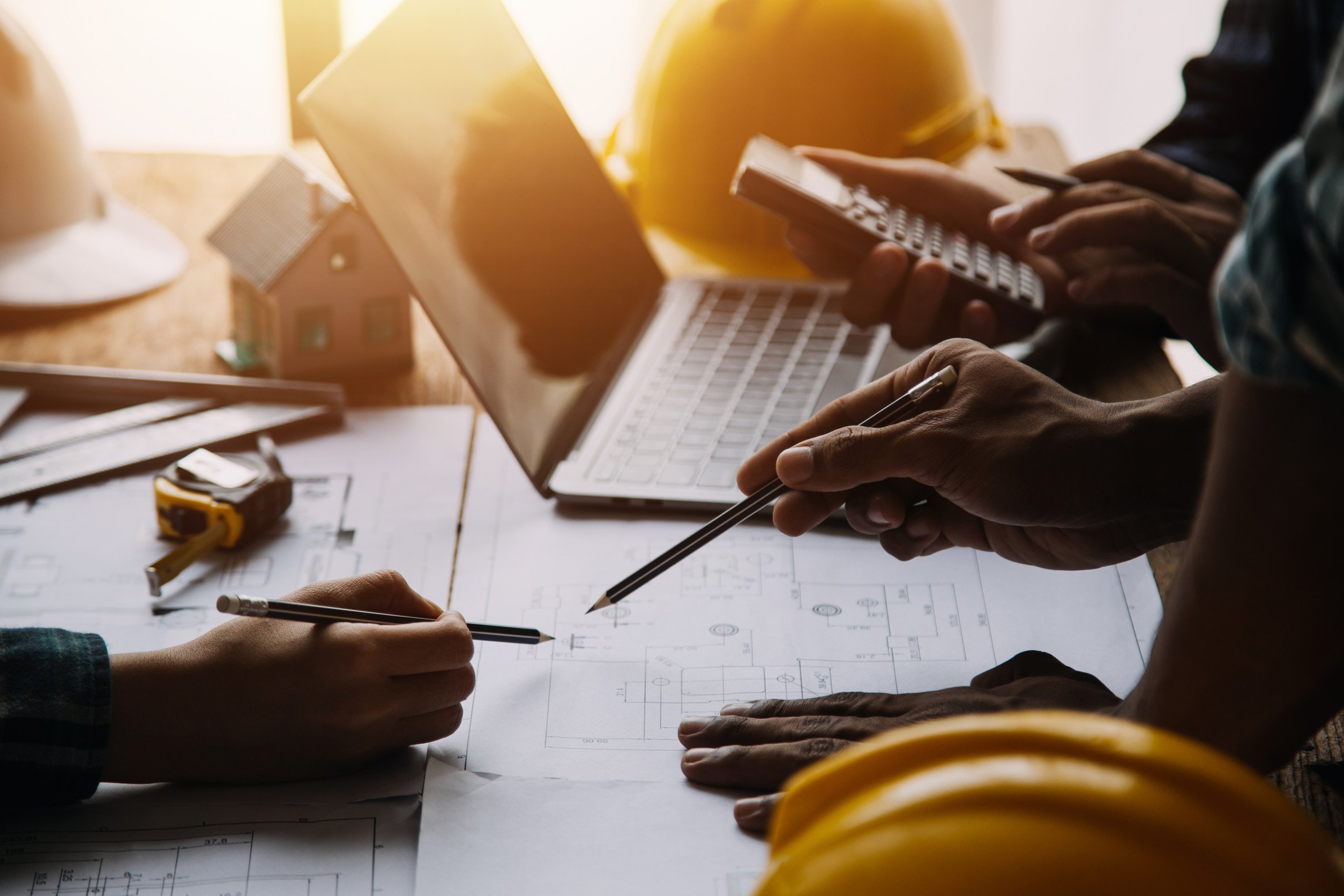 several people gathered around a table covered in blueprints and a laptop, some of them holding pens and calculators, working toward a common goal