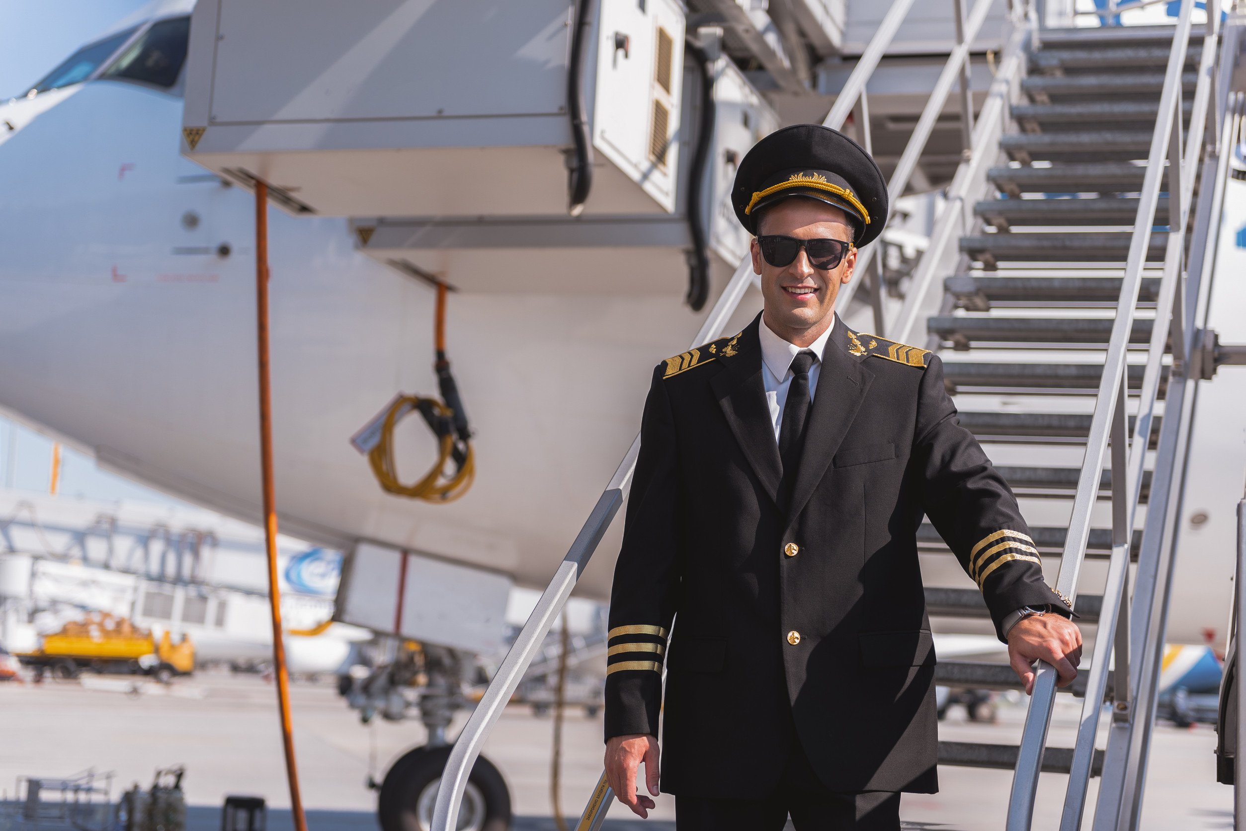 airline pilot in aviator sunglasses standing on a mobile staircase docked on a commercial jet in the background