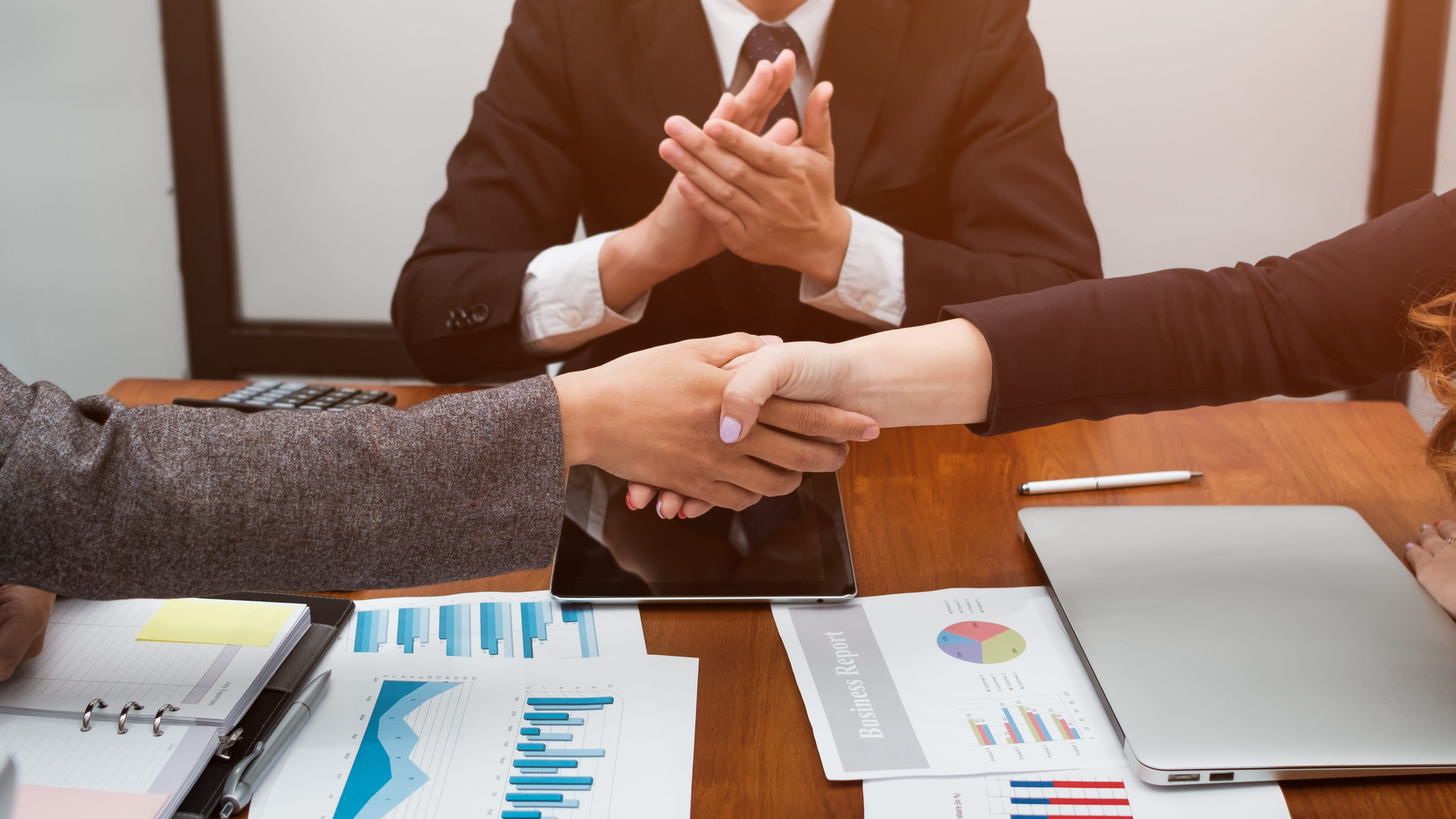 two business professionals shake hands over a table with various charts and graphs as a third person claps in the background