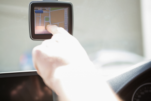 Picture of someone sitting behind the wheel of a car. A screen using GPS is mounted to the windshield, and the person is touching it.