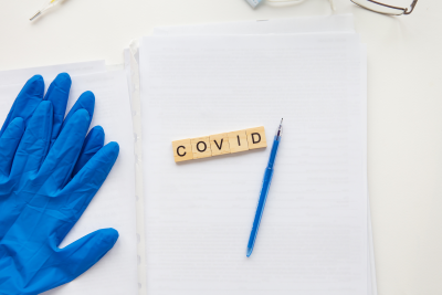 the word 'covid' spelled in scrabble letters atop a stack of papers, flanked by blue gloves and a needle