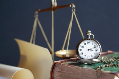 foreground: old fashion pocket watch resting an worn old book, next to a rolled scroll; background: miniature justice scale