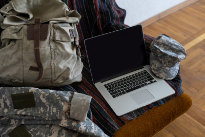 A military uniform, backpack, and camouflage cap are laid out next to an open laptop on a cushioned chair with a striped blanket.