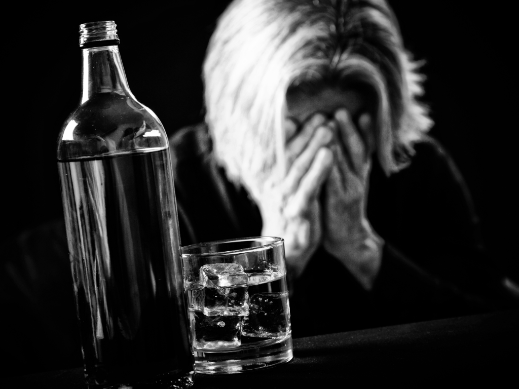 a bottle and a glass of alcohol in the foreground, and a person with their hands on their face in an expression of despair in the background