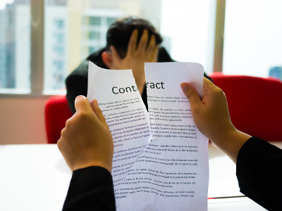 two hands ripping apart a contract in the foreground as a man sitting at a table holds his head in one hand in the backgroud