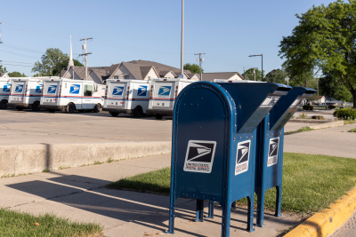 USPS blue mail boxes on side of road in front of row of mail trucks.