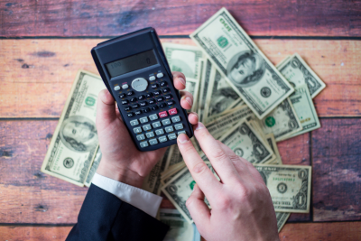 hands of a person operating a calculator over a desk covered in dollar bills spread haphazardly