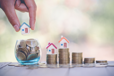 A hand places a small model house into a glass jar filled with coins, next to three stacks of coins topped with miniature houses