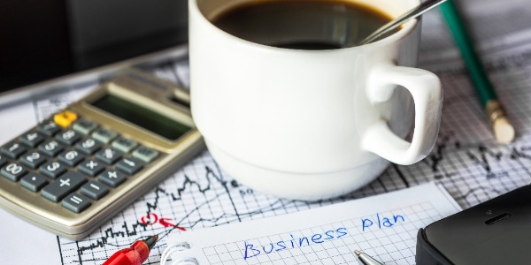 various office supplies including a notebook, calculator, pens and pencils, financial graphs and a cup of coffee laid out on a desk